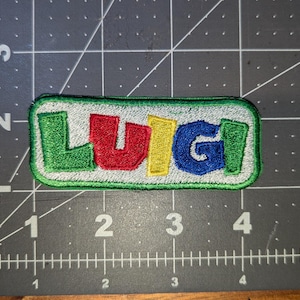 May include: Embroidered patch featuring the name "LUIGI" in colorful letters. The letters are green, red, yellow, and blue, with a white border and a green outline. The patch is rectangular and sits on a dark gray surface.