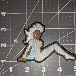 May include: An embroidered patch featuring a white and gray figure with yellow accents. The figure is sitting with one leg extended and the other bent. The figure has a brown face and white hair.