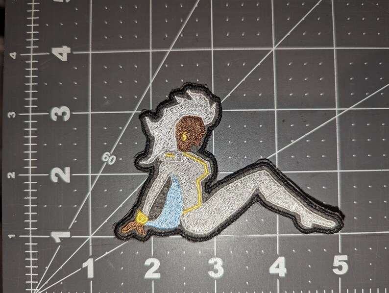 May include: An embroidered patch featuring a white and gray figure with yellow accents. The figure is sitting with one leg extended and the other bent. The figure has a brown face and white hair.