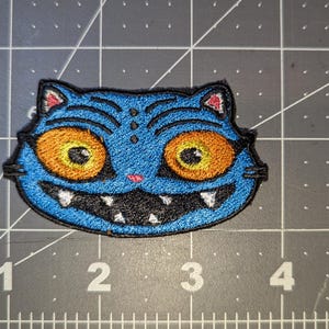 Peut inclure: Écusson brodé représentant un visage de chat bleu avec de grands yeux orange et un large sourire. Le chat a des moustaches noires et des accents rouges sur les oreilles. L'écusson mesure environ 5 cm de large.