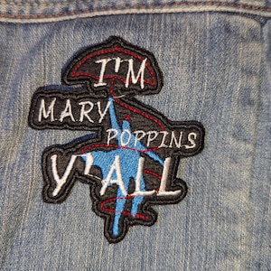 May include: Embroidered patch with the text "I'M MARY POPPINS Y'ALL" in white lettering on a black background. The patch features a blue silhouette of Mary Poppins with a red umbrella and red lines. The patch is sewn onto a denim jacket.