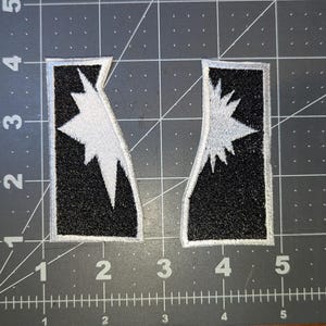 May include: Two black and white embroidered patches. Each patch has a black background with a white starburst design. The patches are rectangular with a silver border. The patches are placed on a gray cutting mat.
