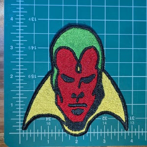 May include: Embroidered patch of Vision from Marvel Comics. The patch shows Vision's face in red, green, and yellow, with black outlines. It is placed on a green cutting mat, with measurements in centimetres.