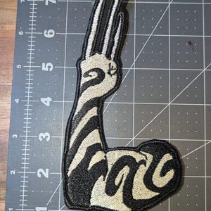 May include: Embroidered patch featuring a stylized arm with a hand gesture. The design uses black and off-white colors, with a zebra-like pattern on the arm and hand. The patch is likely for clothing or accessories.