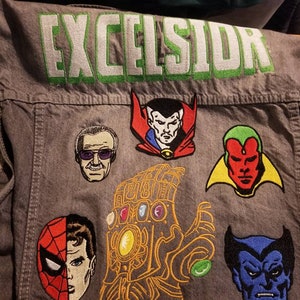 May include: Grey denim vest featuring embroidered Marvel character patches, such as Spider-Man, Doctor Strange, and Vision. The word "EXCELSIOR" is embroidered across the top in green and white.