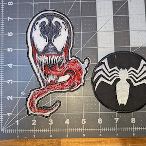 May include: Two embroidered patches featuring Venom. One patch is a white and black Venom face with a red tongue. The other is a black circular patch with a white spider symbol.