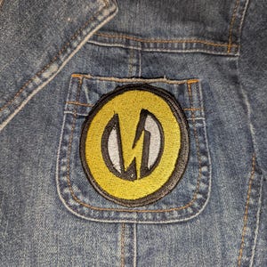 May include: Close-up of a denim jacket with a yellow and black patch on the pocket. The patch features a white and gray lightning bolt design inside a yellow circle. The jacket is blue with visible stitching.