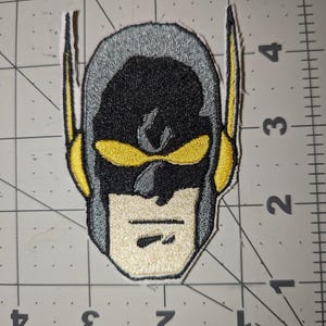May include: Embroidered patch featuring a superhero helmet design. The patch has a black face and a gray helmet with yellow accents. The patch is on a gray and white grid background.
