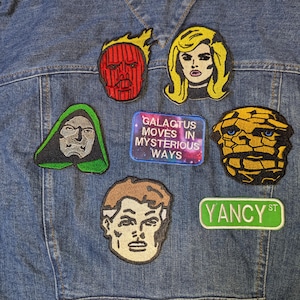 May include: A denim jacket adorned with several embroidered patches featuring comic book characters and phrases. Patches include a red-faced figure, a blonde woman, a green-hooded figure, a rocky-skinned character, a brown-haired face, and a patch that reads "GALACTUS MOVES IN MYSTERIOUS WAYS."