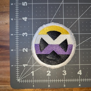 May include: Round embroidered patch featuring the non-binary pride flag colors: yellow, white, purple, black, and gray. The design includes a stylized X shape in the center, surrounded by a white border. The patch is likely for clothing or accessories.