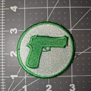 May include: A round patch featuring a green handgun design on a gray background, encircled by a green border. The patch is likely an embroidered emblem, suitable for application on clothing or accessories.