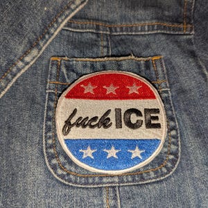 May include: Close-up of a denim jacket with a patch on the pocket. The patch is round with a red and white top section, a gray center, and a blue bottom section. The text "fuck ICE" is embroidered on the patch.