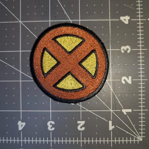May include: Embroidered patch with a circular design. It has a brown background and a yellow 'X' shape in the center, with a black border. The patch is on a gray cutting mat with centimeter measurements.