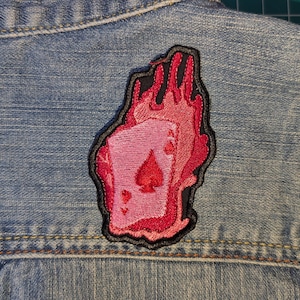 May include: A denim vest with a decorative patch. The patch features a pink Ace of Spades card design engulfed in red and black flames. The patch is embroidered with a black border.