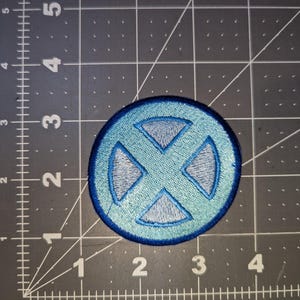 May include: A round, embroidered patch featuring a light blue and gray design. The patch has a dark blue border and a stylized "X" symbol in the center. The patch is likely for clothing or accessories.