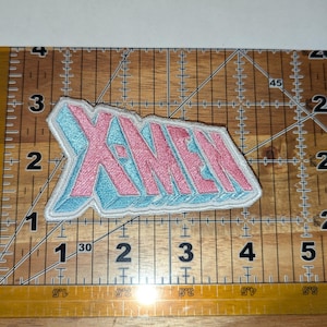 May include: Embroidered X-MEN patch with pink lettering and a light blue outline. The patch is on a clear ruler with inch measurements, and the background is a wooden surface.