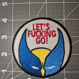 Peut inclure: Écusson brodé de forme circulaire. Il présente un motif bleu et jaune, avec le texte "LET'S FUCKING GO!" en rouge. L'écusson est bordé d'un contour noir et d'un fond gris.