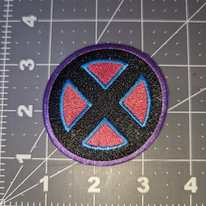 May include: Circular embroidered patch with a black background and a red and blue X-shaped design. The patch has a purple border and is placed on a gray cutting mat.