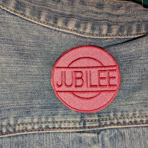 May include: A circular, embroidered patch in a vibrant pink hue featuring the word JUBILEE stitched in capital letters across the center. The patch is securely attached to a light blue denim fabric with visible stitching details.