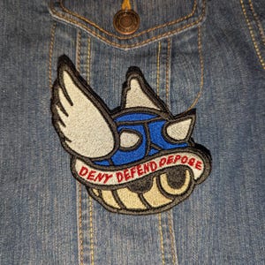 May include: Embroidered patch featuring a blue and white winged design with the words "DENY DEFEND DEPOSE" in red. The patch is sewn onto a denim jacket.