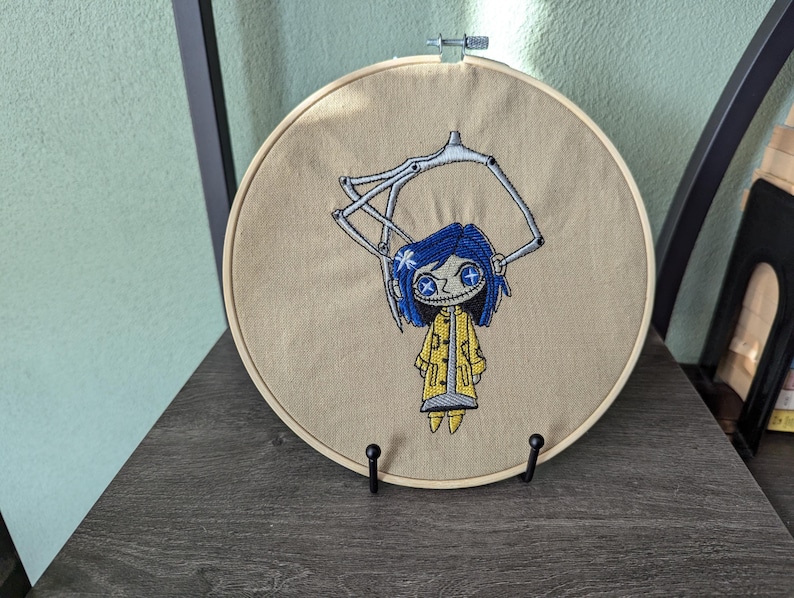 May include: Embroidered artwork featuring a character with blue hair, a yellow coat, and button eyes. The design is set within a wooden embroidery hoop. The artwork is displayed on a dark wooden surface.