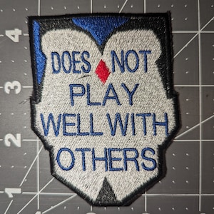 Peut inclure: &Eacute;cusson brod&eacute; blanc avec bordure noire et bleue. L'&eacute;cusson dit "Does not play well with others" avec un diamant rouge au milieu.