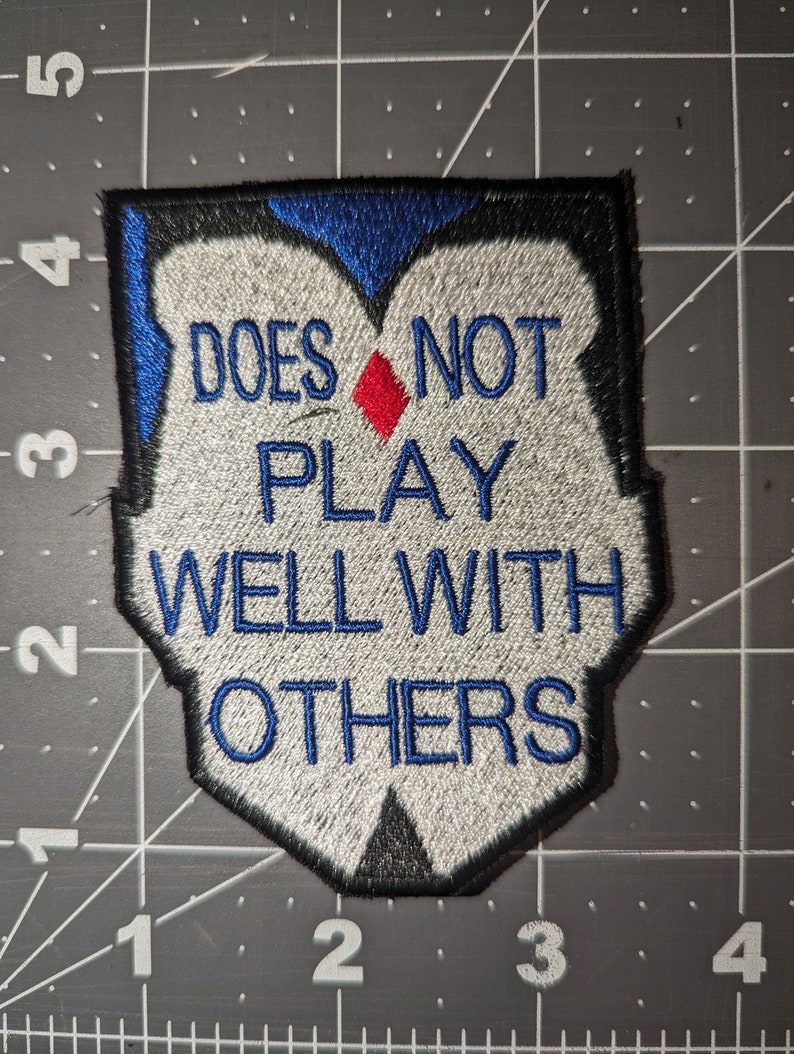 Peut inclure: &Eacute;cusson brod&eacute; blanc avec bordure noire et bleue. L'&eacute;cusson dit "Does not play well with others" avec un diamant rouge au milieu.