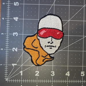 May include: Embroidered patch featuring a stylized face with a white head, red visor, and orange detail. The patch is on a gray cutting mat with a grid pattern, perfect for jackets, bags, or other fabric items.