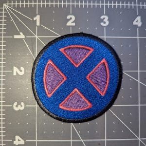 May include: A circular embroidered patch featuring a blue background with a purple "X" design. The "X" is outlined in red. The patch has a black border and is likely for clothing or accessories.
