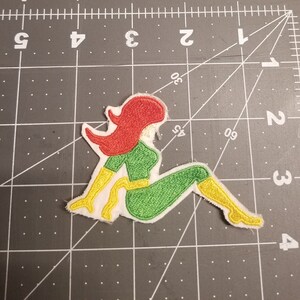 May include: A green and yellow embroidered patch of a woman sitting with red hair.
