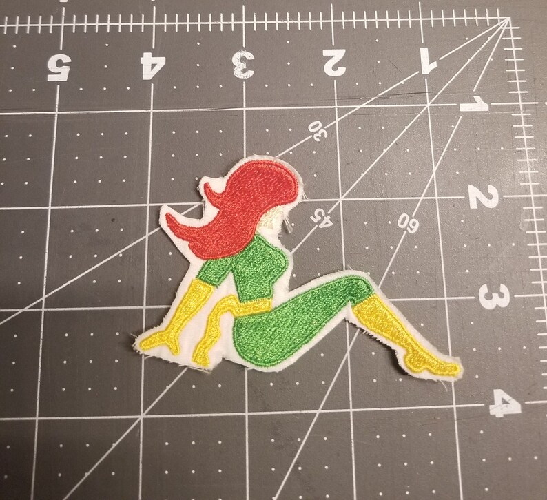 May include: A green and yellow embroidered patch of a woman sitting with red hair.
