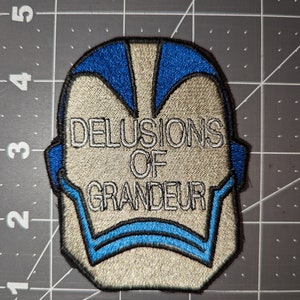 Peut inclure: Un &eacute;cusson brod&eacute; blanc et bleu avec le texte "DELUSIONS OF GRANDEUR" en blanc.