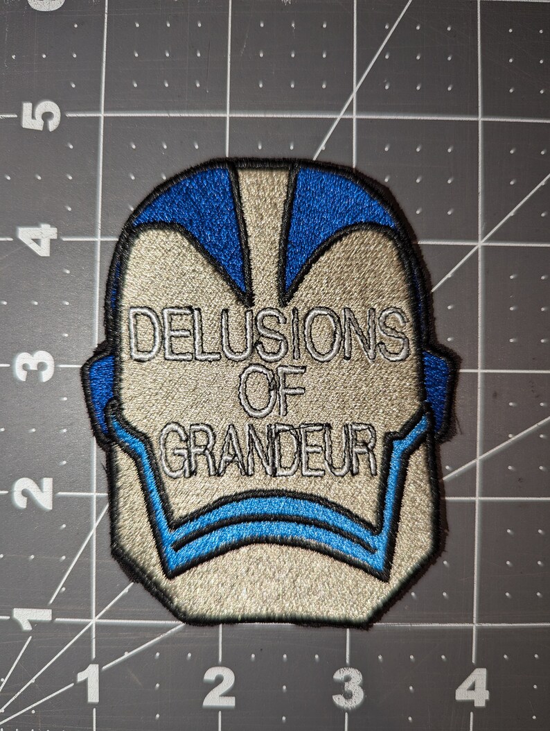 Peut inclure: Un &eacute;cusson brod&eacute; blanc et bleu avec le texte "DELUSIONS OF GRANDEUR" en blanc.