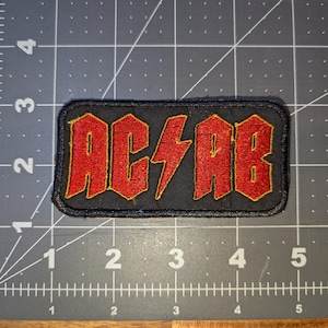 May include: Black rectangular patch with the red letters "AC/AB" and a lightning bolt in the center. The letters have a gold outline. The patch is on a gray surface with measurement markings.