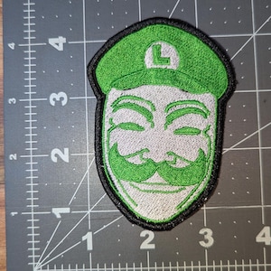 May include: Embroidered patch featuring a green hat with a white "L" and a stylized face with a green mustache and a smiling expression. The patch has a black border and is on a gray surface with measurement markings.