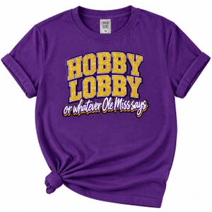May include: A purple short-sleeve t-shirt with a crew neckline and rolled sleeves. The front features the yellow text HOBBY LOBBY in a block font, followed by the white script text or whatever Ole Miss says, with a purple and yellow drop shadow.
