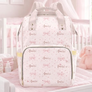 Puede incluir: Un bolso para pañales rosa y blanco con un estampado de lazos y corazones, con el nombre "Amiri" impreso. El bolso tiene un asa superior, correas para los hombros y múltiples bolsillos. Un conejito de peluche rosa está cerca.