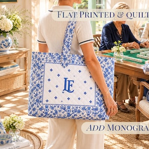 May include: A quilted tote bag featuring a white center panel with a blue monogram of the letters LF, surrounded by a blue and white floral pattern. The bag has a rectangular shape and a wide shoulder strap. Text on the image reads FLAT PRINTED & QUILTED and ADD MONOGRAM.
