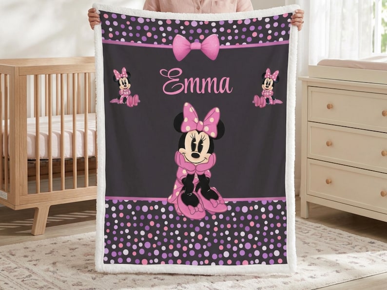 May include: A rectangular fleece blanket featuring a dark gray center with a large Minnie Mouse graphic, the name Emma in pink script, and a pink bow. The top and bottom borders have a black background with pink and purple polka dots.