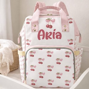 May include: A white and light pink backpack featuring a repeating pattern of red cherries with pink bows. The name Aria is printed in large, bold pink letters on the front panel. The bag includes top handles, adjustable shoulder straps, and a front pocket.