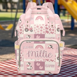 Può includere: Borsa per pannolini patchwork rosa e bianca a tema fattoria. La borsa presenta un motivo ripetuto di mucche, trattori, arcobaleni e fiori. Il nome "Millie" è scritto sulla tasca anteriore.