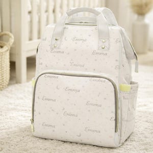 May include: A white baby diaper bag with the name "Emma" printed repeatedly, along with stars and crescent moons. The bag has a front pocket, side pockets, and a top handle. The bag is on a white carpet.