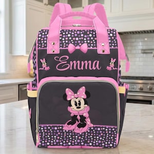 May include: A black and pink backpack featuring a Minnie Mouse design. The backpack has a front pocket with a Minnie Mouse graphic, the name "Emma", and polka dot accents. It has pink straps and handles.