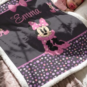 May include: A soft, plush blanket featuring a dark gray background with a Minnie Mouse graphic, pink polka dot borders, and a large pink bow. The name Emma is printed in a cursive pink font. The blanket has a white sherpa fleece trim.