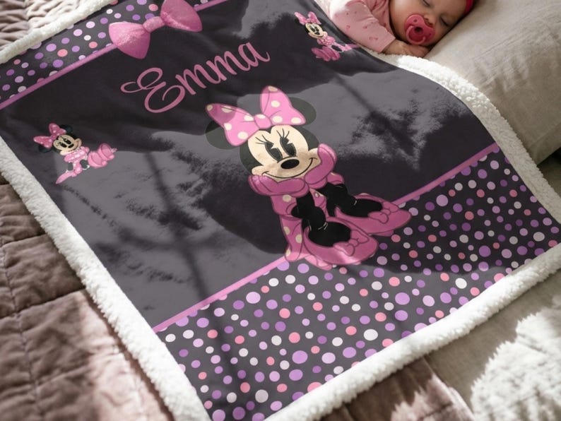 May include: A soft, plush blanket featuring a dark gray background with a Minnie Mouse graphic, pink polka dot borders, and a large pink bow. The name Emma is printed in a cursive pink font. The blanket has a white sherpa fleece trim.