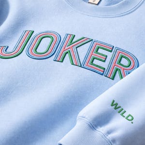May include: Light blue sweatshirt with the word "JOKER" embroidered in green, pink, and blue. The sleeve features the word "WILD." in green. The sweatshirt is made of a soft material.