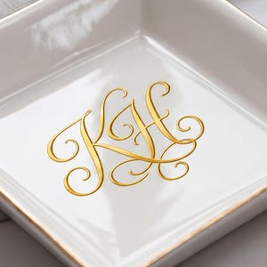 May include: A square white ceramic trinket dish with a gold-trimmed rim. The center of the dish features a gold-colored monogram with the letters KH in a decorative, swirling script font. The surface of the dish is smooth and glossy.