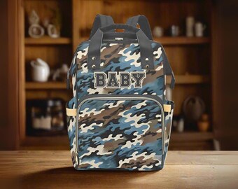 Puede incluir: Una mochila con un patrón de camuflaje en azul, marrón y blanco, con la palabra BABY impresa en una fuente oscura de bloque en el bolsillo frontal. La bolsa incluye correas gris oscuro, un asa superior y bolsillos laterales con ribetes claros.