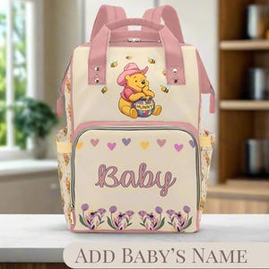 Puede incluir: Una bolsa de pañales Winnie the Pooh rosa y crema con un diseño floral y la palabra "Baby" impresa en la parte delantera. La bolsa presenta una imagen de dibujos animados de Winnie the Pooh sosteniendo un tarro de miel.
