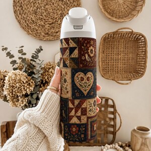 May include: A cylindrical water bottle featuring a multi-colored patchwork quilt pattern. The design includes squares with floral motifs, geometric shapes, birds, and a central heart emblem in shades of navy blue, deep red, cream, and olive green.